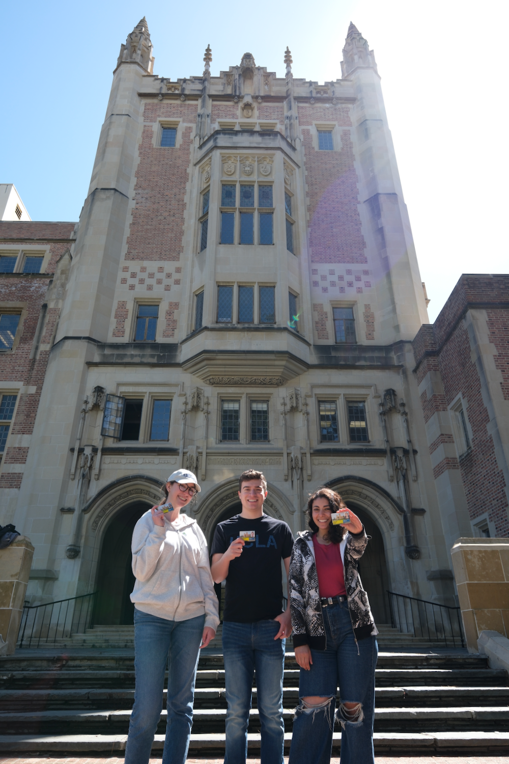Three students standing outside of Kerkhoff hall holding out their BruinCards